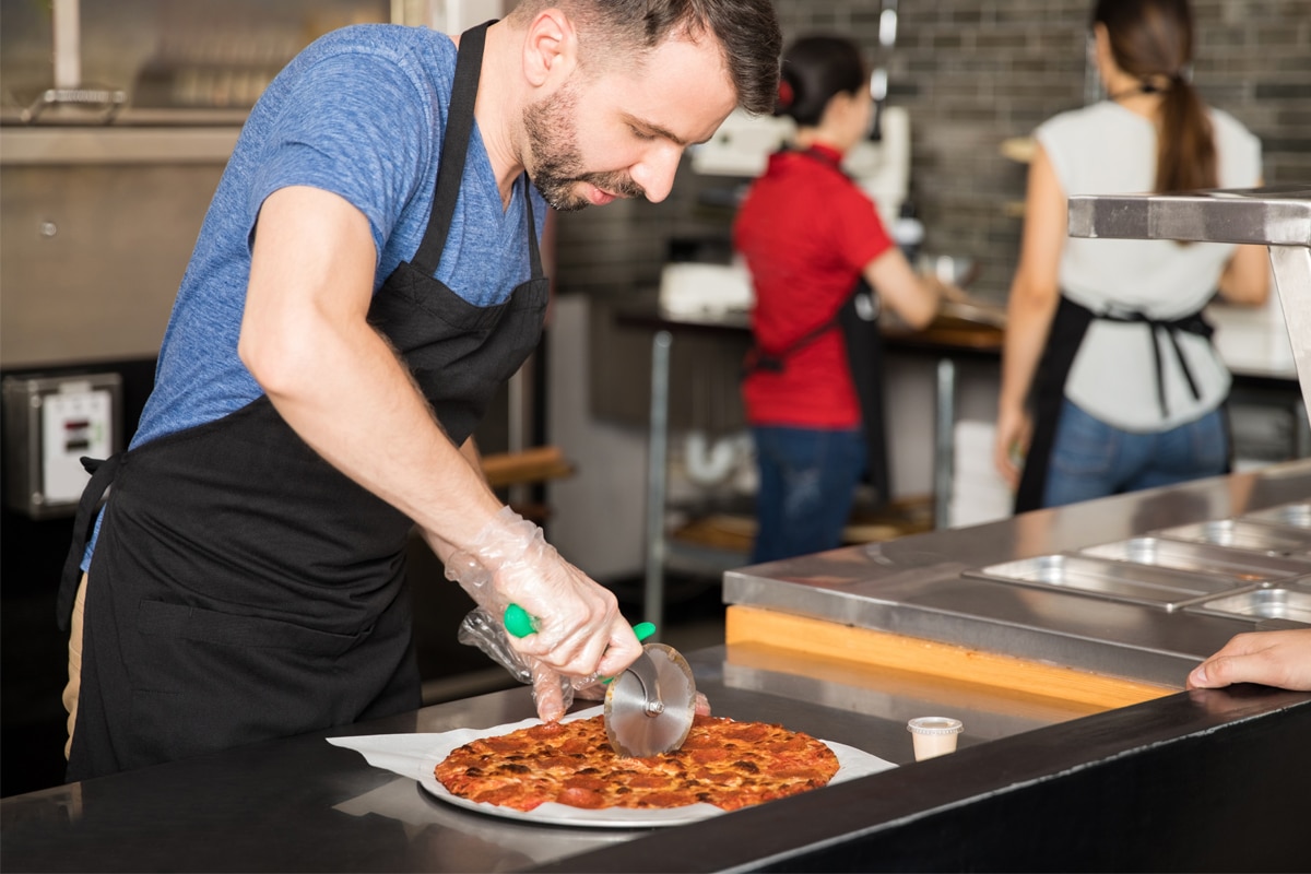 Man in blue shirt and apron cutting a pizza for delivery.